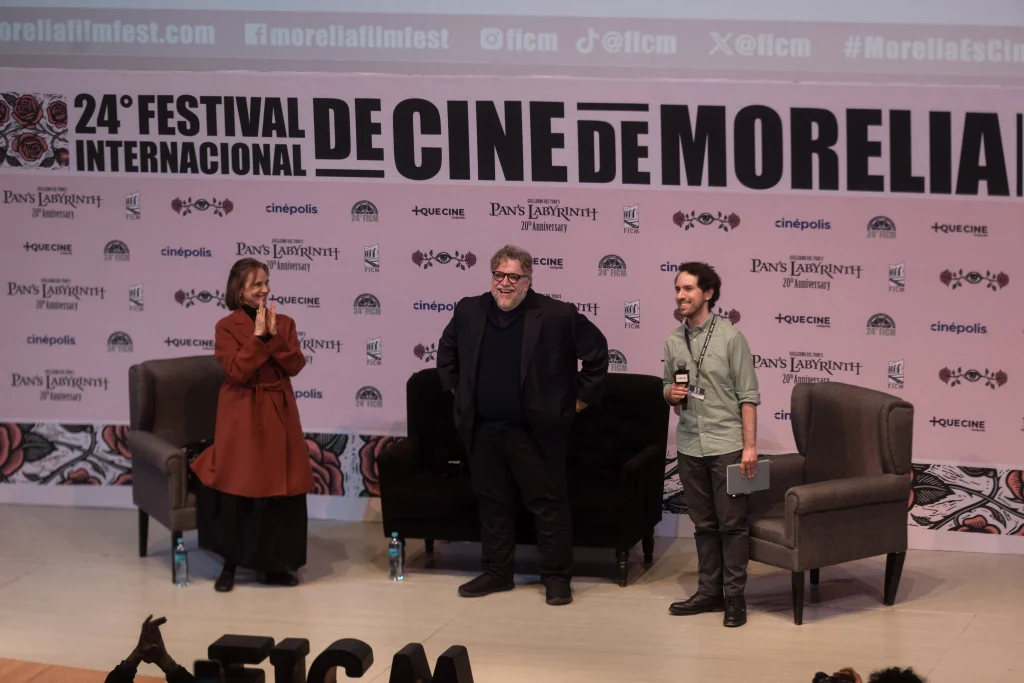 Guillermo del Toro sonríe sobre el escenario acompañado de dos participantes durante la clase magistral del Festival Internacional de Cine de Morelia 2026.
