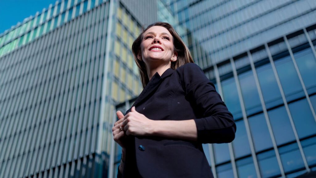 Mujer emprendedora sonriendo frente a edificios modernos de oficinas, como representación del liderazgo femenino en los negocios.