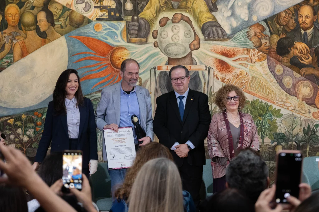 Juan Villoro sostiene el diploma y el galardón del Premio Carlos Fuentes 2026, acompañado por tres representantes institucionales frente a un mural de Diego Rivera.