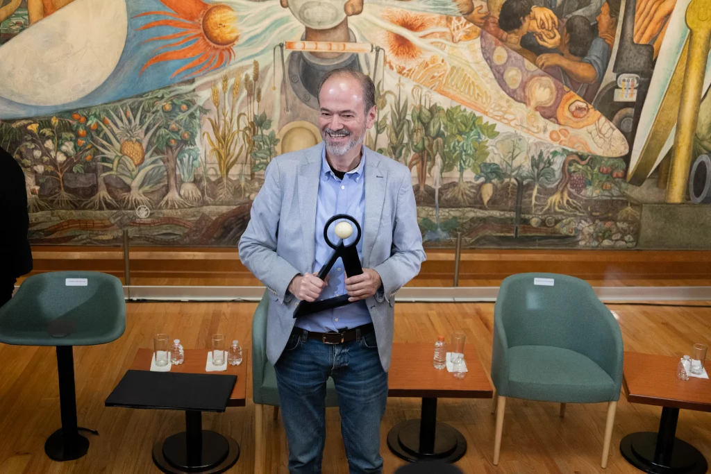 Juan Villoro sonríe mientras sostiene el Premio Carlos Fuentes 2026 frente a un mural en el Palacio de Bellas Artes.